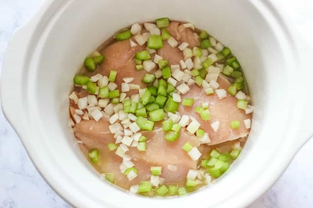 Raw chicken breasts in a crockpot with diced onions, celery and chicken stock on top.