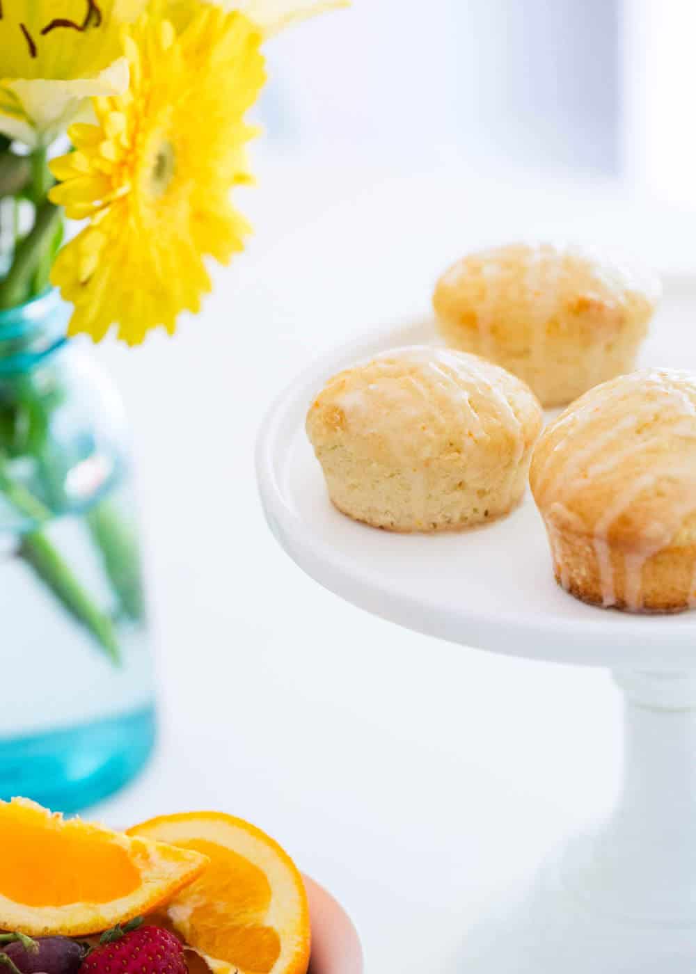 Orange muffins on a cake stand next to a vase of yellow flowers.