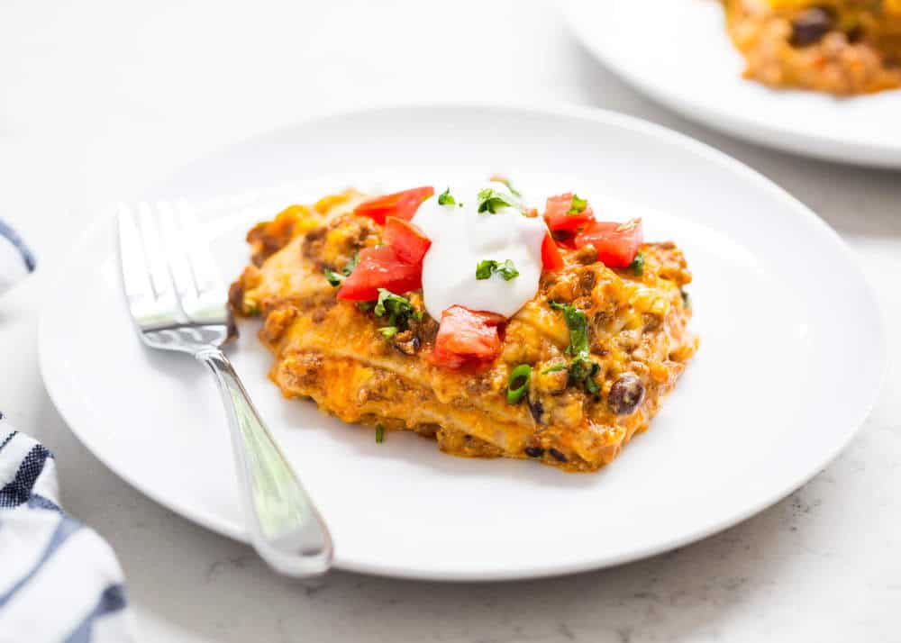 Piece of taco lasagna on a white plate topped with diced tomatoes and sour cream.