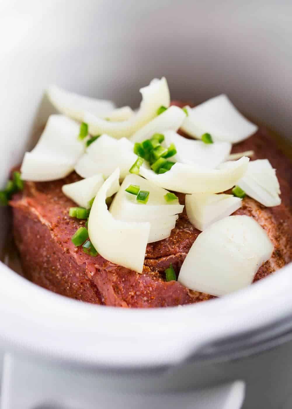 Pork in the slow cooker with chopped onion and jalapeños on top.