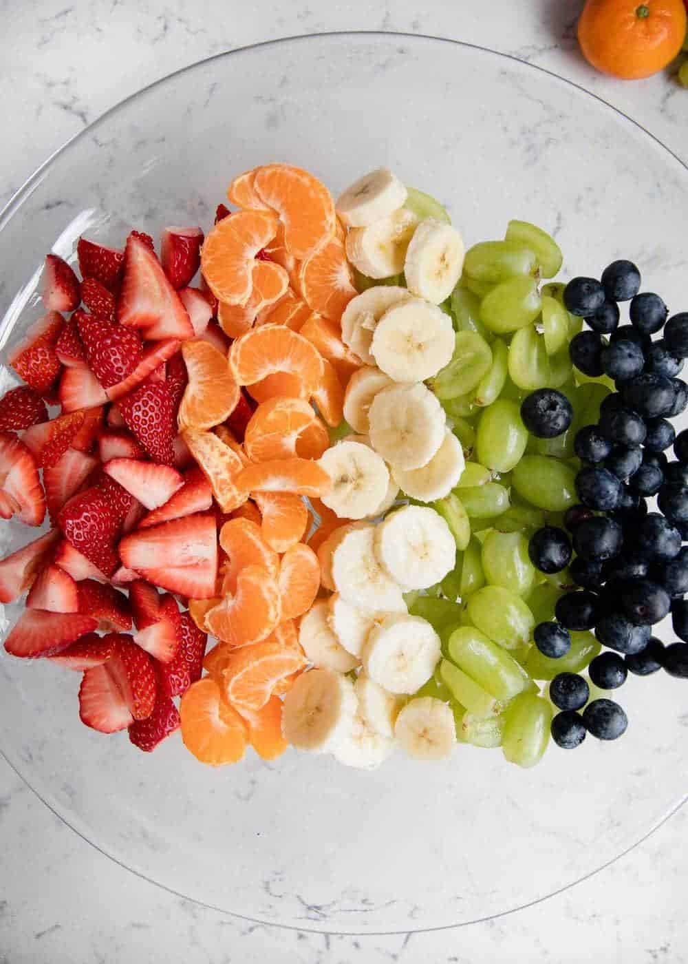 Strawberries, oranges, bananas, grapes, and blueberries are lined up on a glass plate. 