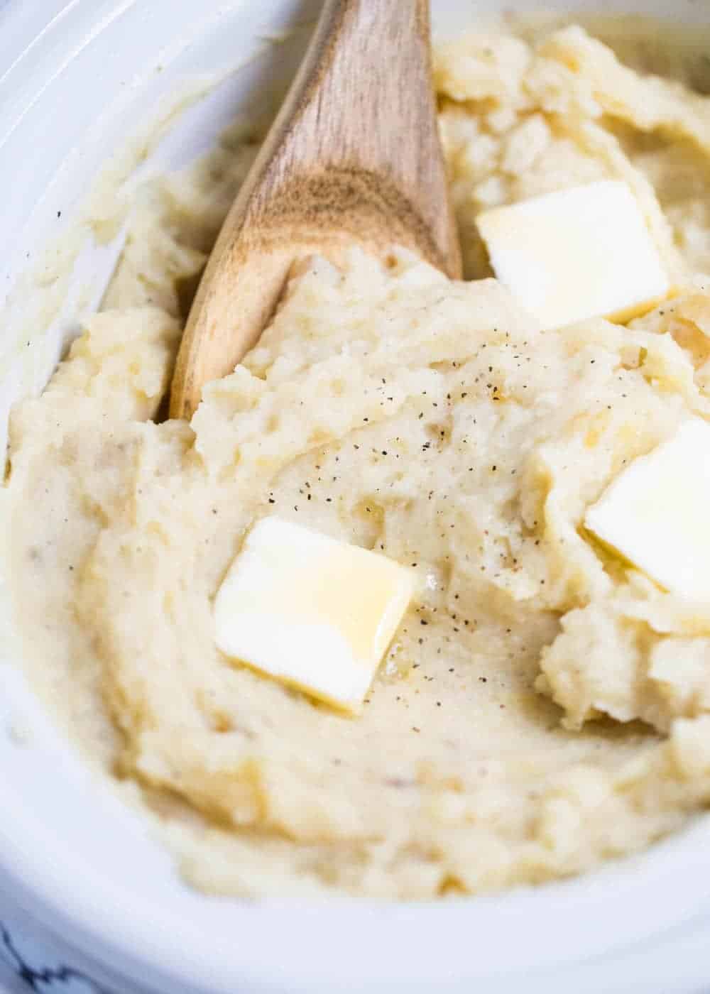 Wooden spoon in slow cooker mashed potatoes with butter slices on top.