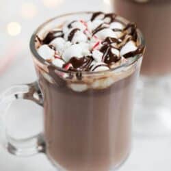 Homemade hot chocolate in a glass mug topped with mini marshmallows, hot fudge and crushed peppermint