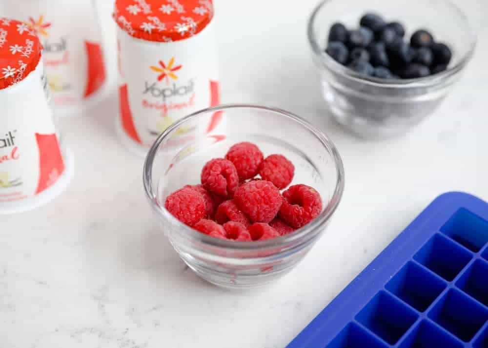 Bowls of raspberries and blueberries on the counter with yogurt containers.