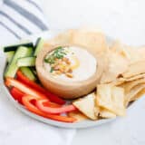 bowl of white bean hummus on a plate with fresh sliced veggies and pita chips