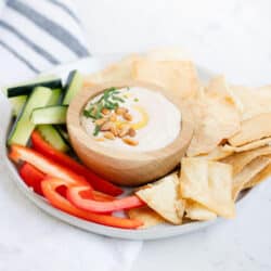 bowl of white bean hummus on a plate with fresh sliced veggies and pita chips