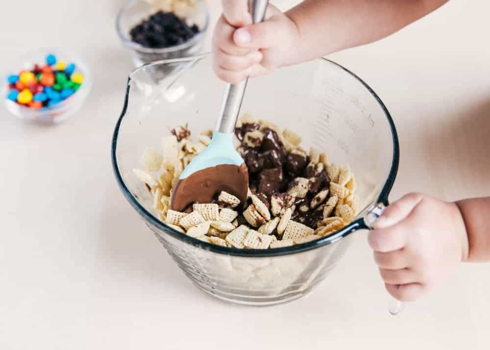Toddler mixing Chex cereal and melted chocolate in a mixing bowl with a spoon.