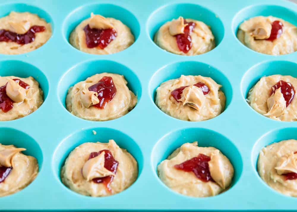 Muffin batter in pan with strawberry jam and peanut butter on top.