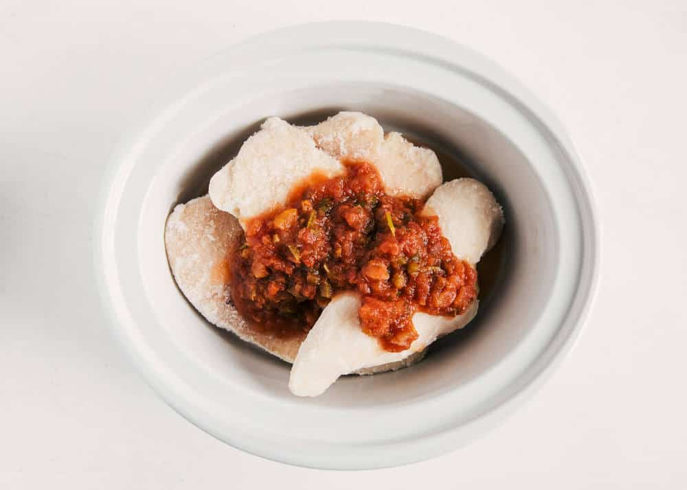 Frozen chicken breast with salsa on top in a white crockpot.