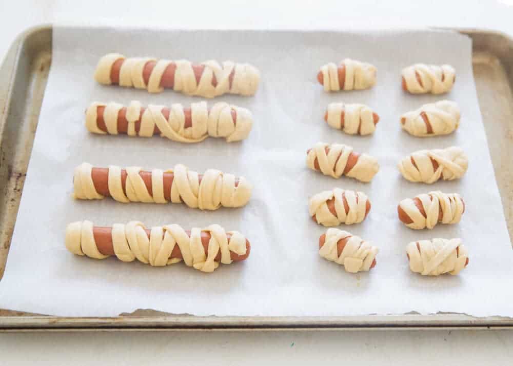 These mummy hot dogs are the easiest Halloween appetizer that takes only 2 ingredients to make! So fun, festive and delicious. Serve with spooky spider web sauce for the ultimate Halloween experience! Mummy hot dogs on baking sheet.