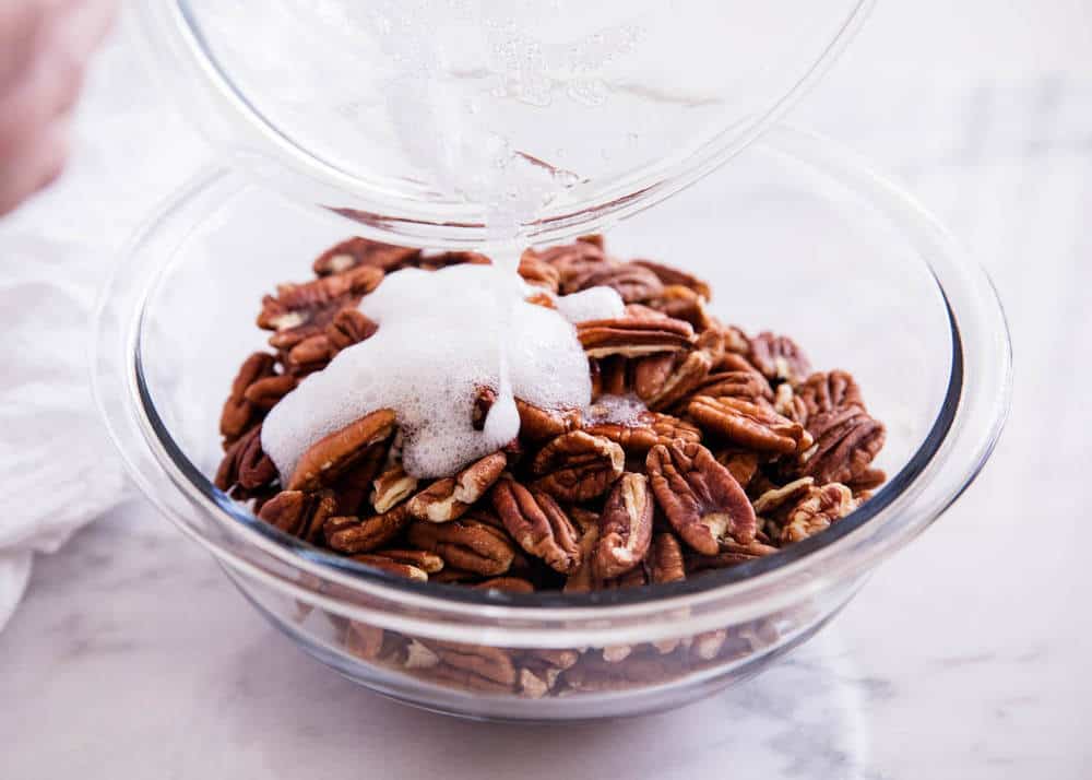 Pouring egg whites over bowl of pecans. 