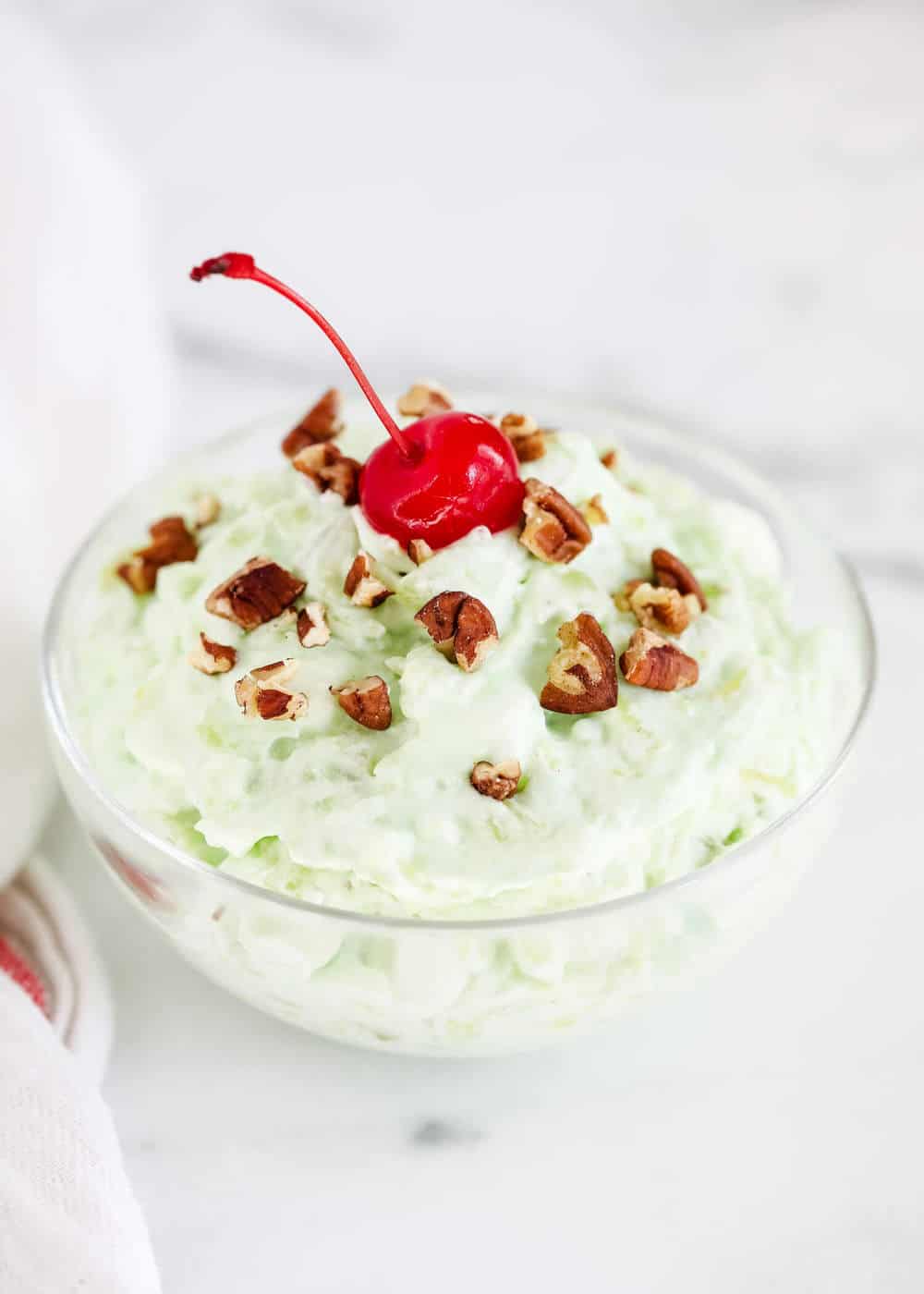 Pistachio pudding salad in a glass bowl with a cherry on top.