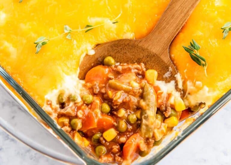 A casserole dish filled with shepherd's pie. The top layer is golden and cheesy, with some of it scooped away to reveal a mixture of ground meat, peas, carrots, corn, and sauce. A wooden spoon rests in the dish. Garnished with fresh herbs.