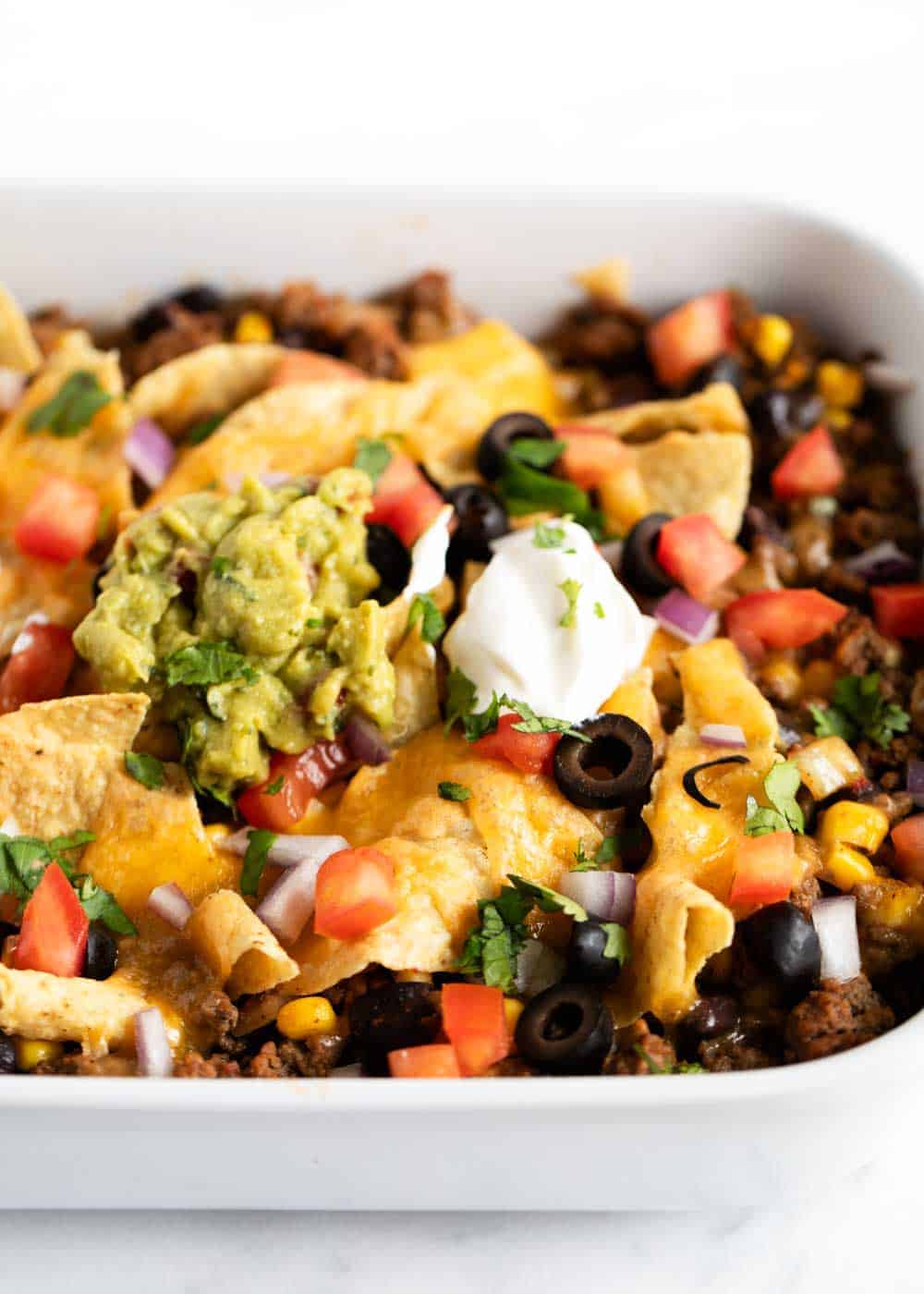 This taco casserole has delicious layers of ground beef, beans, corn, tortilla chips and cheese. Baked to perfection and ready in just 15 minutes. Close up of taco casserole in pan with toppings.