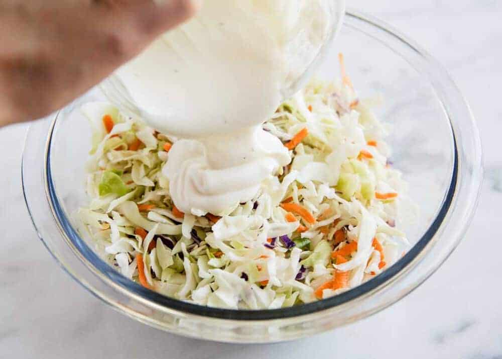 Pouring dressing over coleslaw mix in a glass bowl.