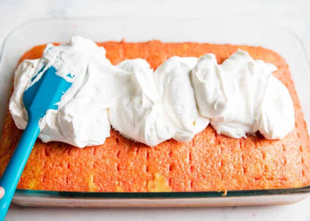 Spreading whipped cream on strawberry jello cake with a spatula.