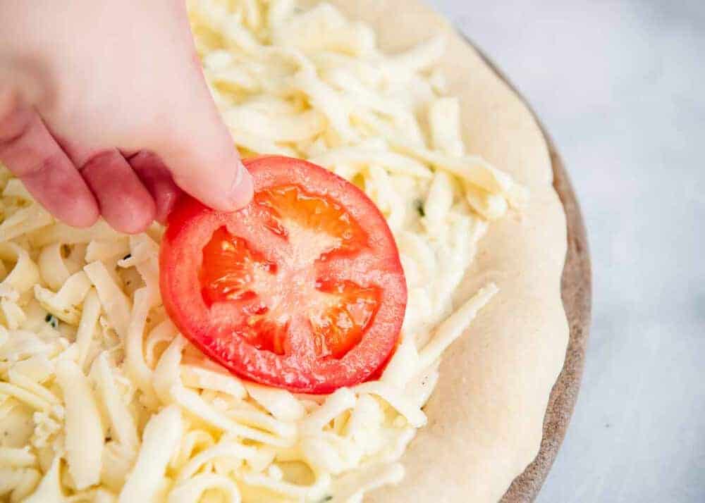 Adding a tomato slice on top of pizza dough with garlic sauce and cheese.