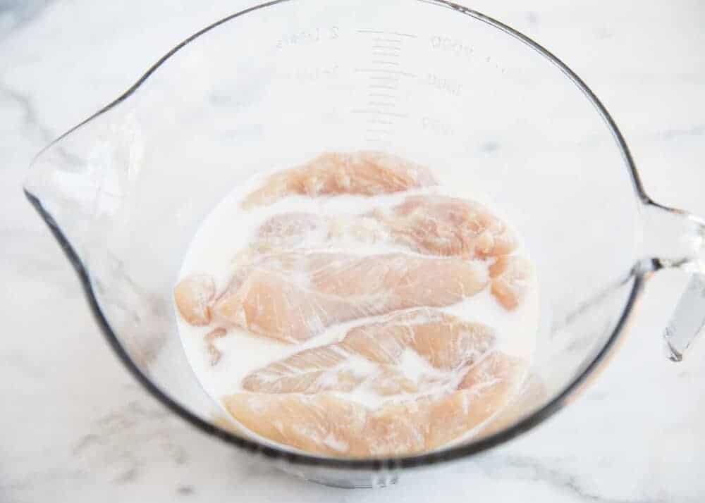 Chicken tenders soaking in buttermilk in glass bowl.