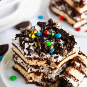 Slice of no-bake ice cream sandwich cake layered with whipped topping, hot fudge, crushed Oreos, and M&Ms on a white plate.