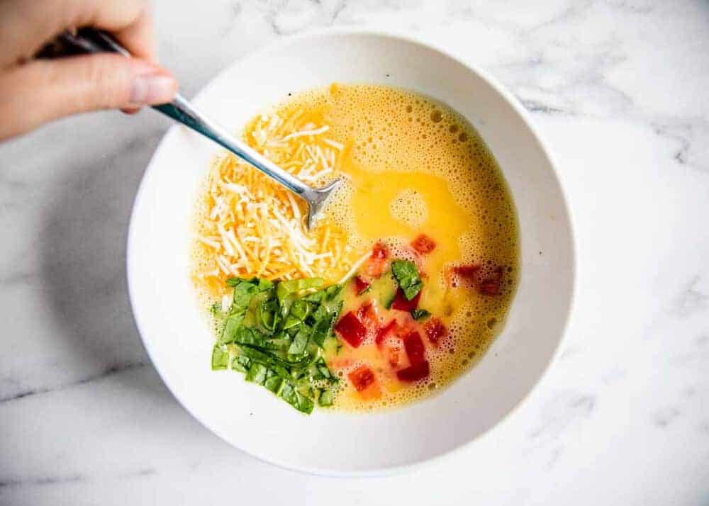 Whisked eggs in a bowl with cheese, spinach and peppers being stirred into the bowl.