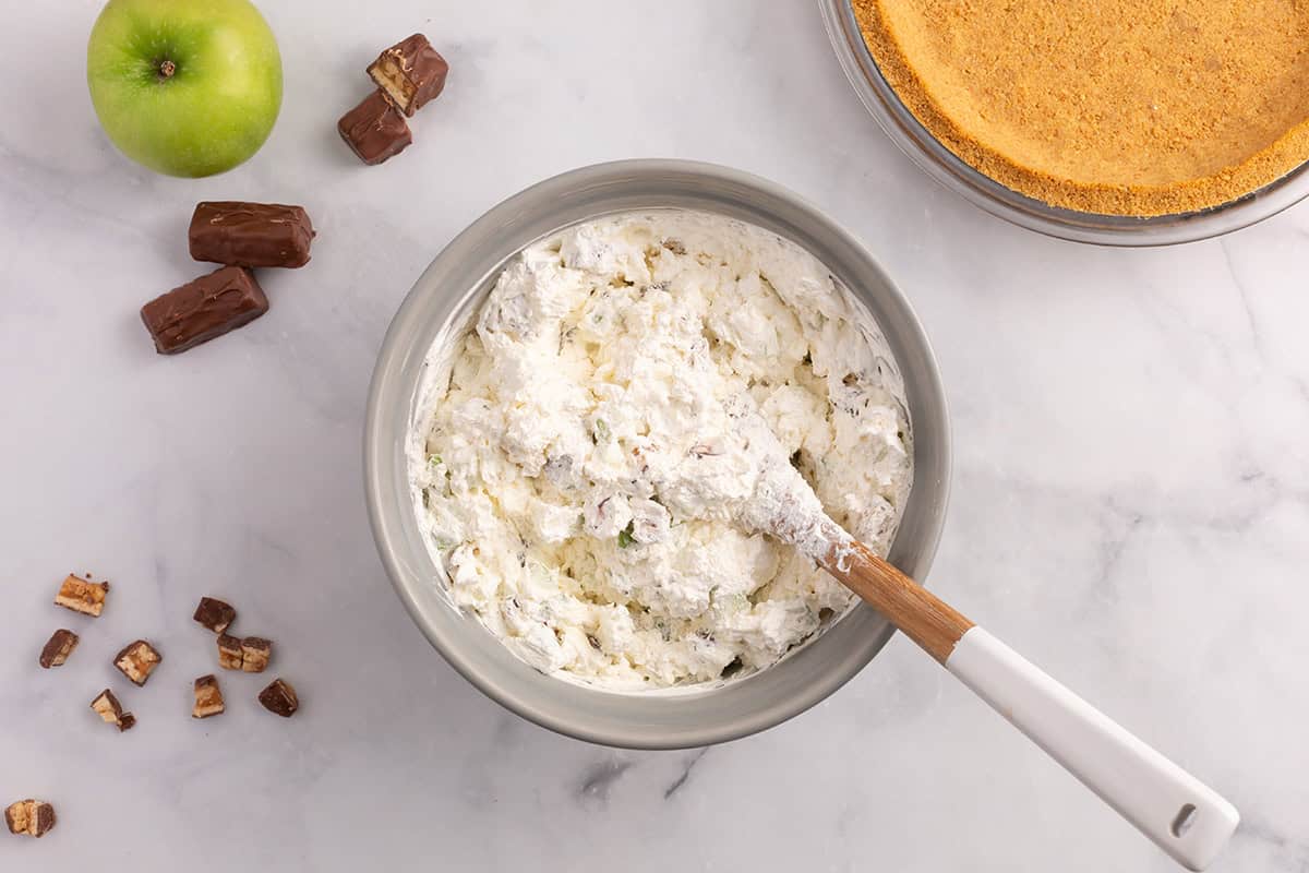 Creamy Snickers apple pie filling mixed in a bowl with chopped apples and candy pieces, ready to add to the graham cracker crust.