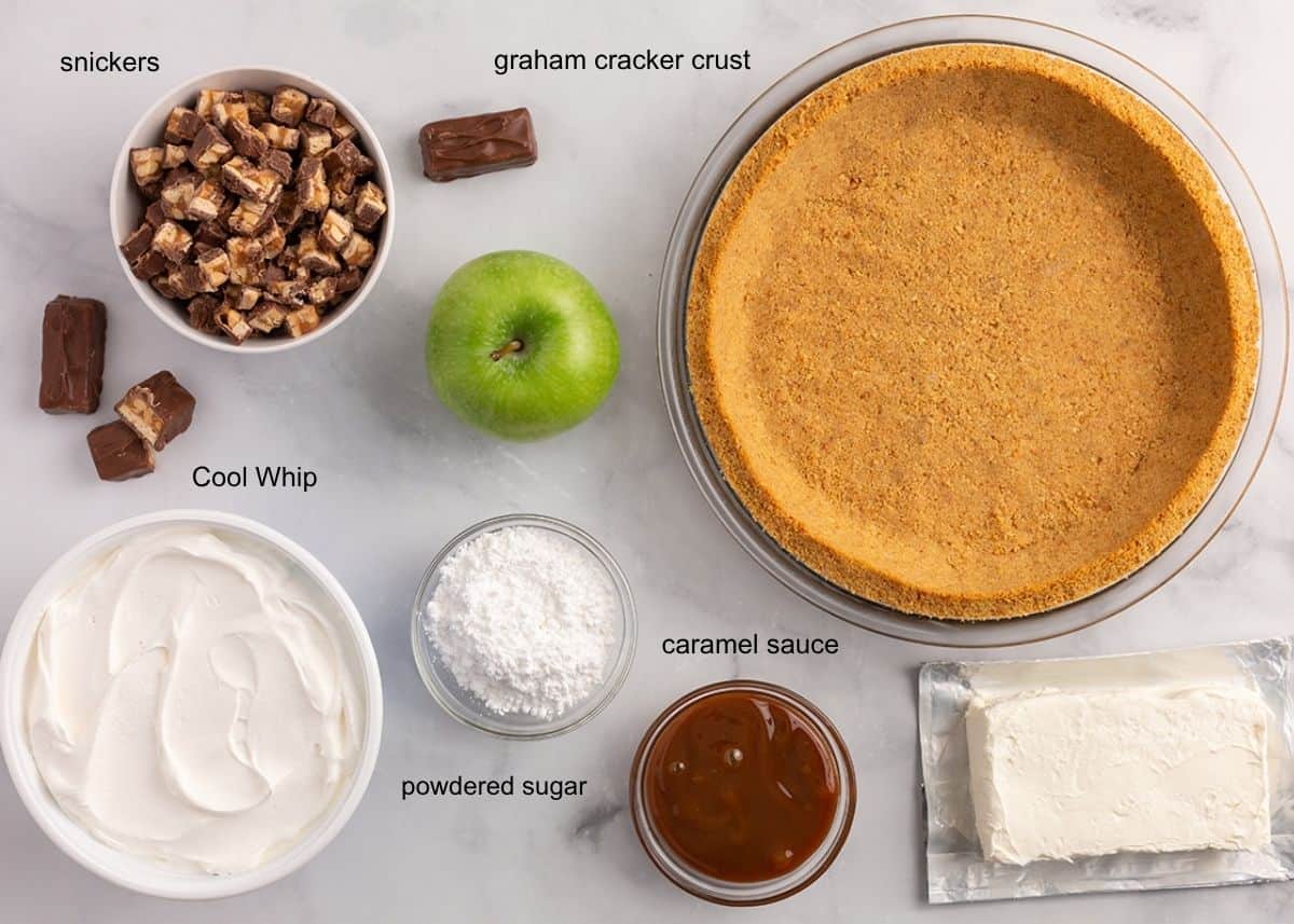Ingredients for Snickers caramel apple pie including chopped Snickers, graham cracker crust, Cool Whip, cream cheese, powdered sugar, caramel sauce and a green apple.