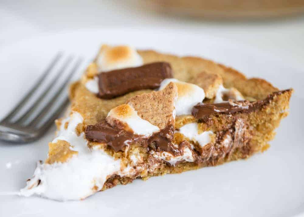 Easy and delicious s'mores pie loaded with gooey marshmallows, chocolate and a graham cracker cookie crust. The perfect summer pie that tastes even better than the traditional campfire s'mores! Piece of s'mores pie on a white plate.