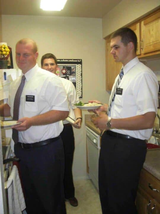 A group of men with plates in their hands, standing in a small kitchen