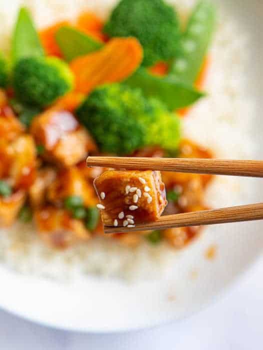 Chopsticks holding piece of chicken from teriyaki bowl. 