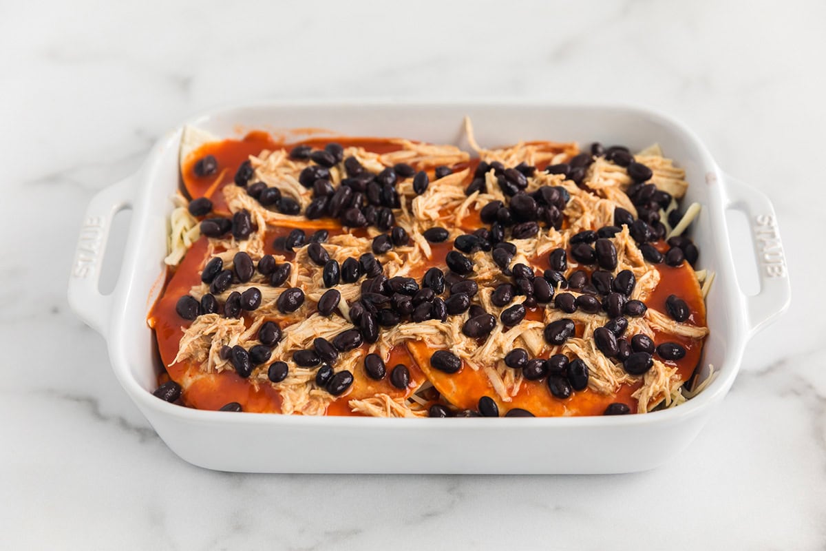 Shredded chicken and black beans topped with red enchilada sauce in a layered chicken enchilada casserole before baking.