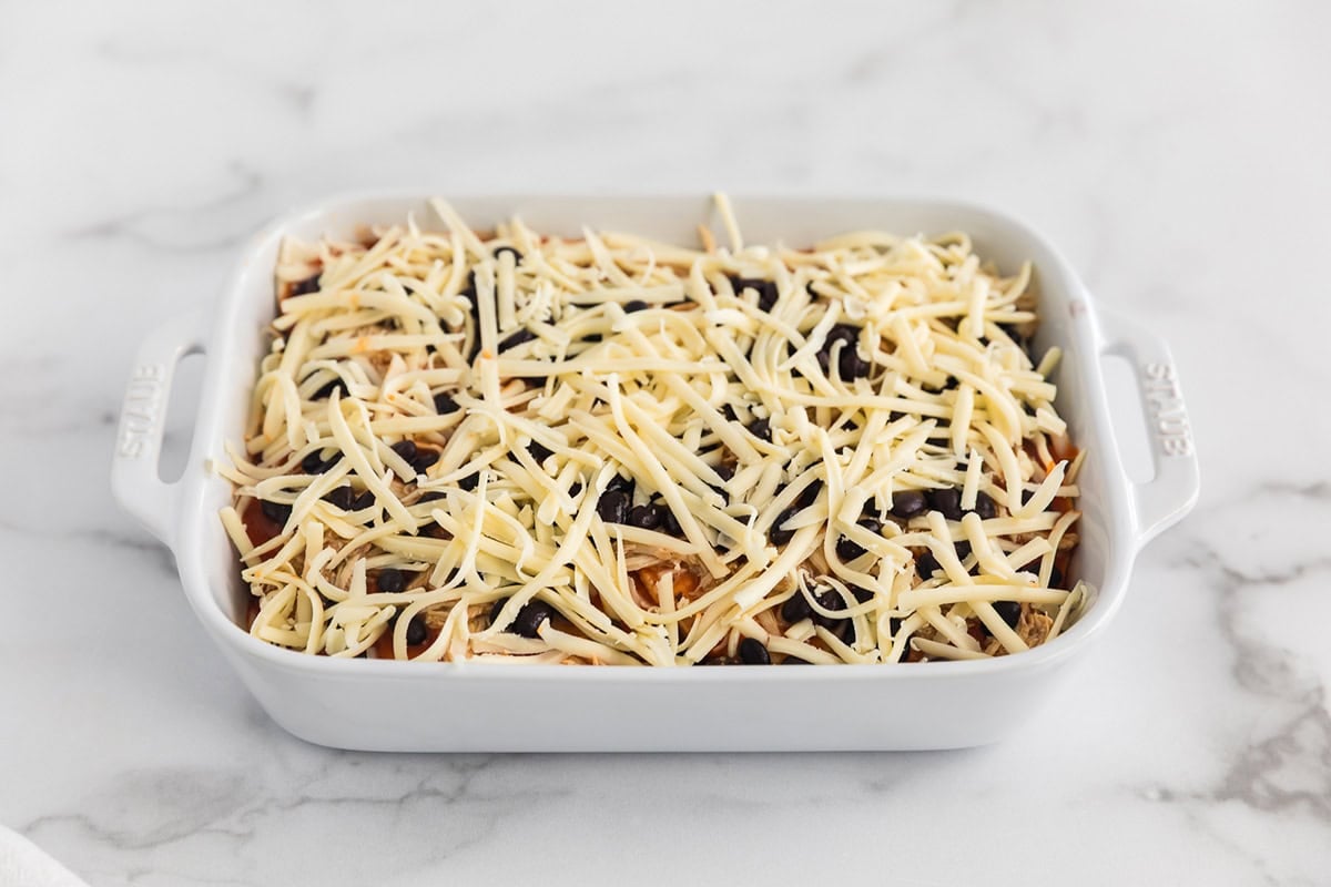 Shredded Monterey Jack cheese sprinkled over chicken, black beans, and enchilada sauce in a layered chicken enchilada casserole.