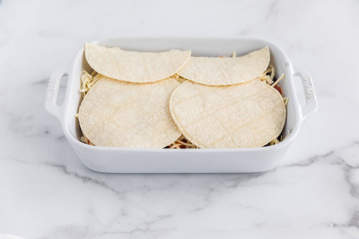 Corn tortillas layered over shredded chicken, black beans, enchilada sauce, and cheese in a white baking dish for chicken enchilada casserole.