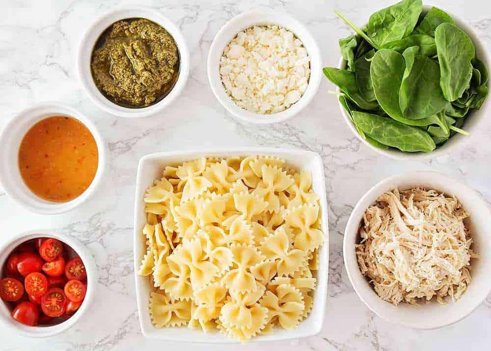 Pesto pasta salad ingredients in bowls on the counter.