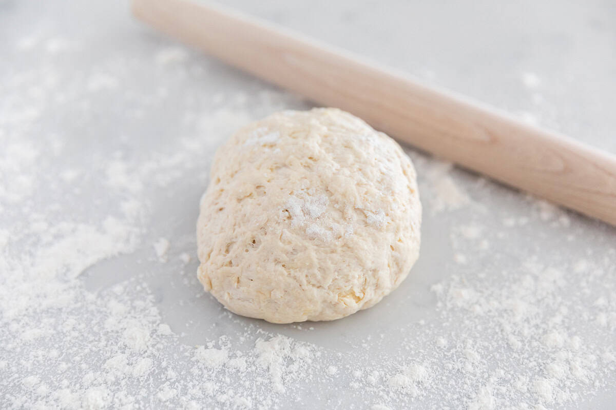 Ball of homemade pizza dough without yeast on a floured surface with a wooden rolling pin.