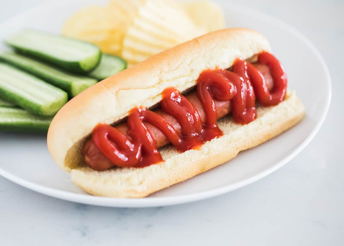 Hot dog on white plate with ketchup.
