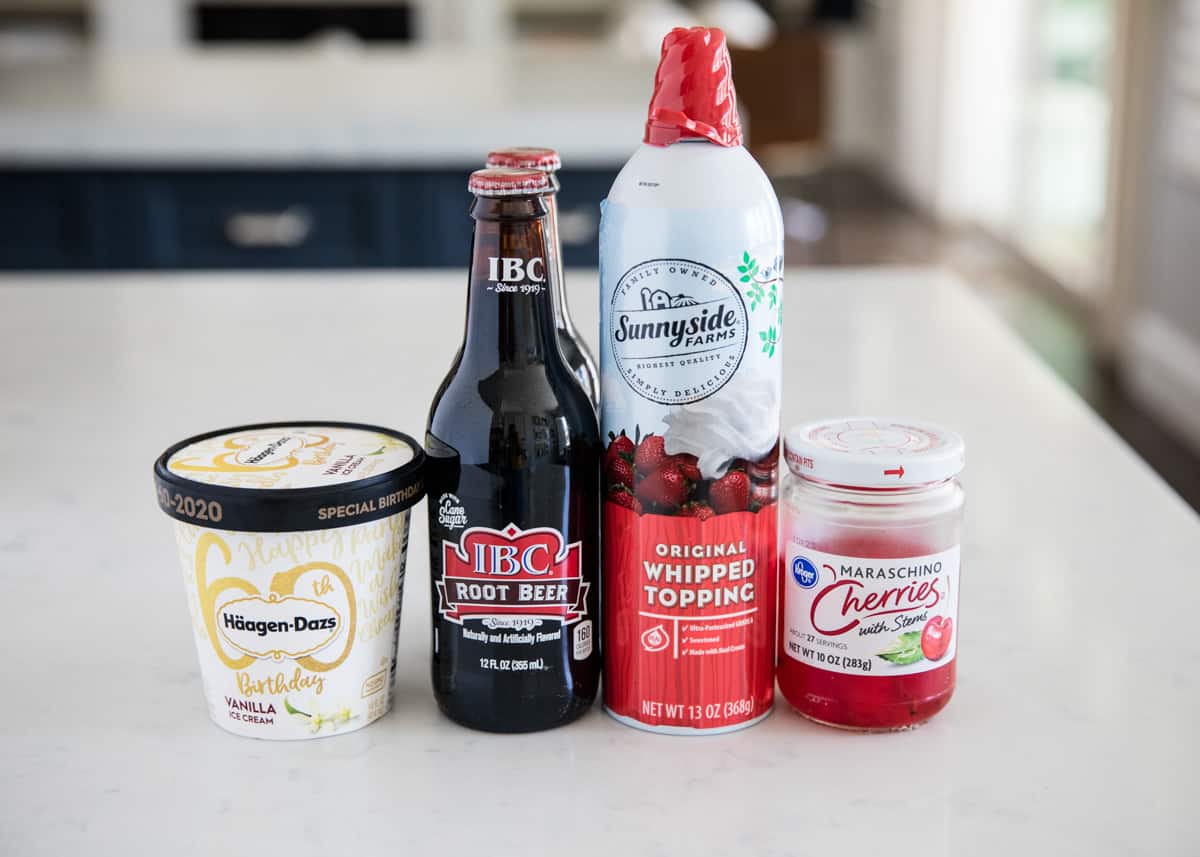 Root beer, ice cream, whipped cream and maraschino cherries on the counter. 