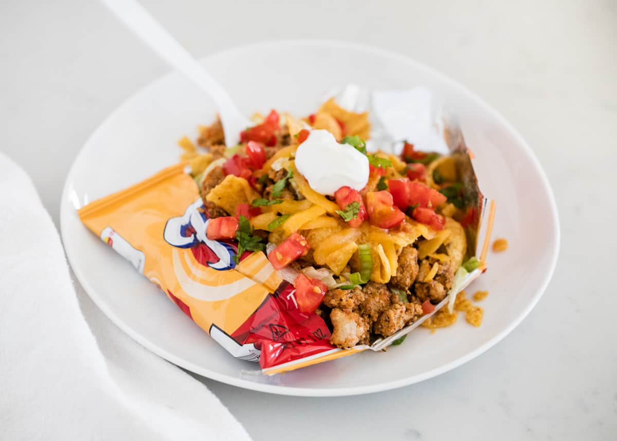 Walking tacos are a simple and delicious dinner idea with hardly any clean up. Your favorite taco meat and toppings served in a bag of Fritos or Doritos. These tacos are perfect for parties and potlucks! Walking taco on white plate.