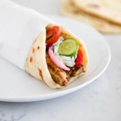 A chicken gyro wrapped in pita bread and served on a white plate with tzatziki sauce and fresh vegetables.