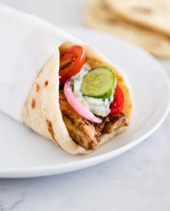 A chicken gyro wrapped in pita bread and served on a white plate with tzatziki sauce and fresh vegetables.
