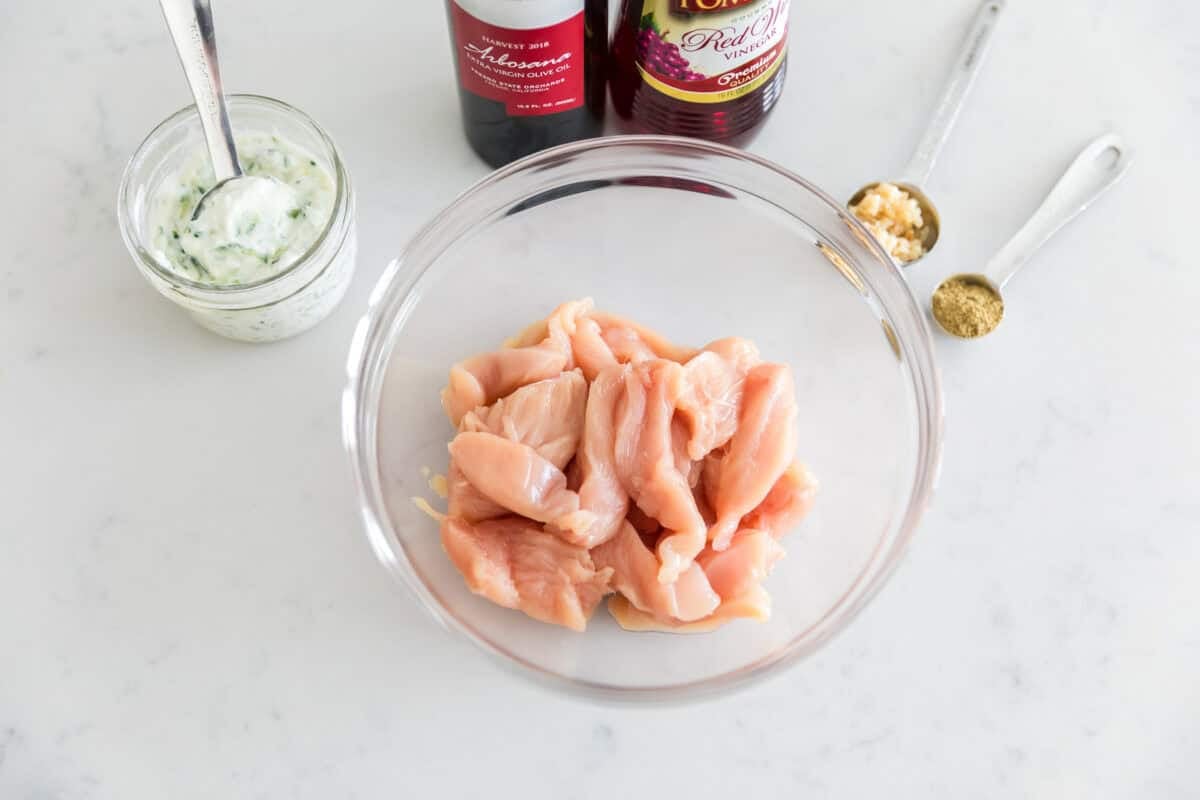 Chicken breast strips in a glass bowl with tzatziki sauce, olive oil, red wine vinegar, garlic and oregano for a chicken gyro marinade to the side.