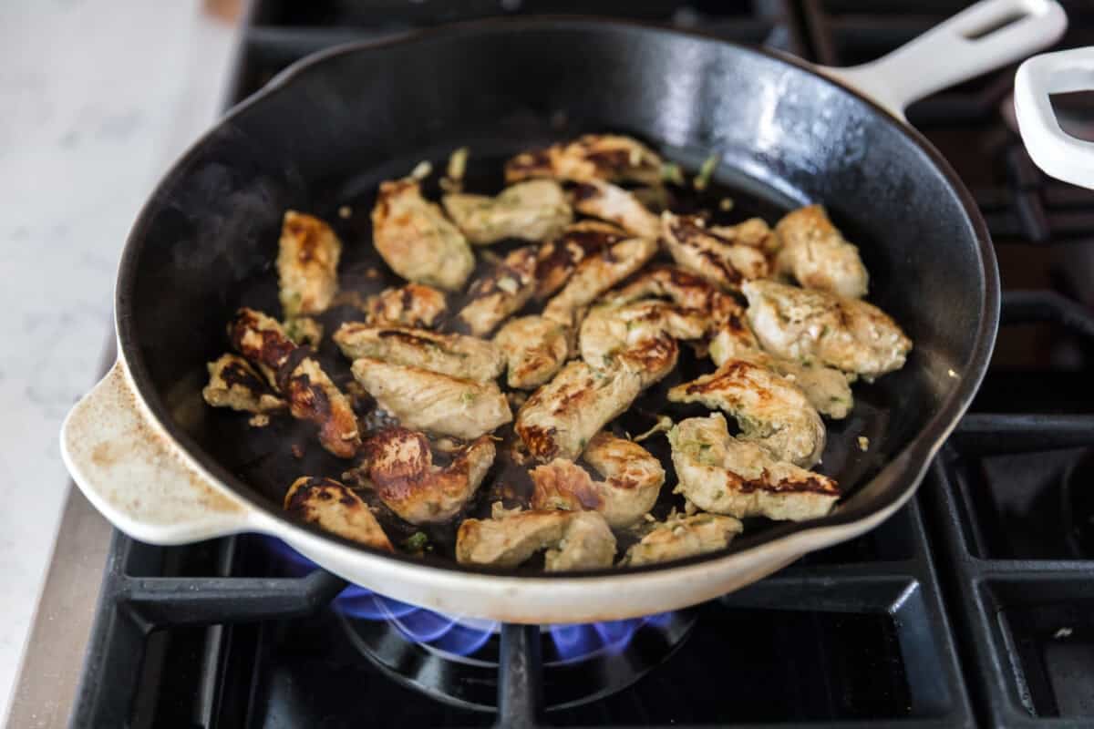 Marinated chicken strips cooking in a cast iron skillet on a gas stovetop.