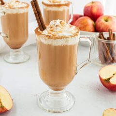 Three glass mugs of caramel apple cider with whipped cream and cinnamon, surrounded by fresh apples and cinnamon sticks.