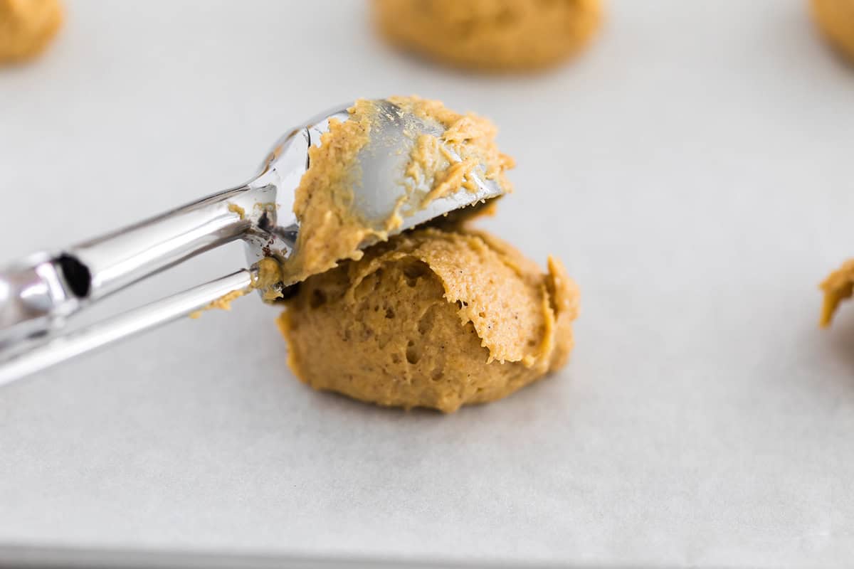 Scooping soft pumpkin cookie dough onto a parchment-lined baking sheet using a medium cookie scoop.