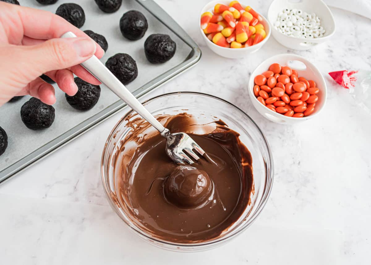 Dipping Oreo truffle in chocolate with candy corn, candy eyes and orange reese pieces in bowls.