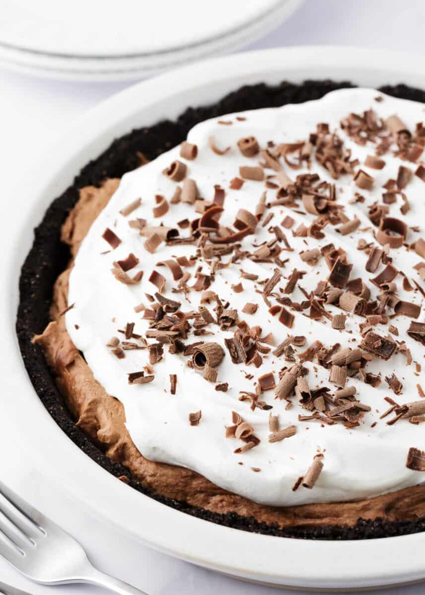 The ultimate French chocolate silk pie made with a chocolate crust, silky smooth chocolate filling and topped with homemade whipped cream and chocolate shavings. This dessert is a chocolate lovers dream! Chocolate silk pie in a white pie dish.