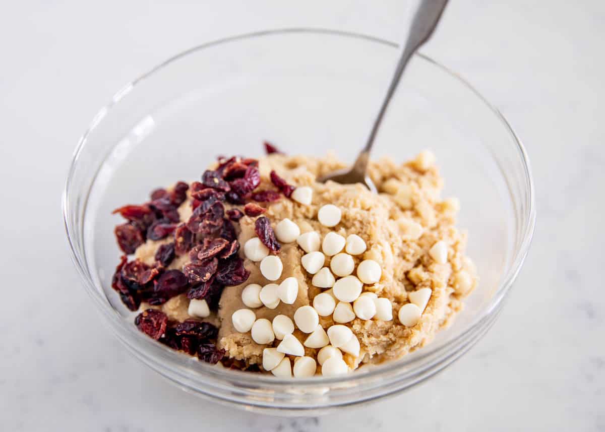 White chocolate chips and dried cranberries being mixed into cookie dough in a glass bowl.