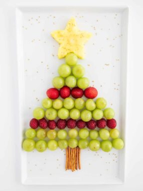A Christmas tree&ndash;shaped fruit platter made with green grapes, strawberries, raspberries, and a pineapple star on top, with pretzel sticks as the trunk.