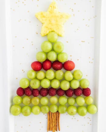 A Christmas tree–shaped fruit platter made with green grapes, strawberries, raspberries, and a pineapple star on top, with pretzel sticks as the trunk.