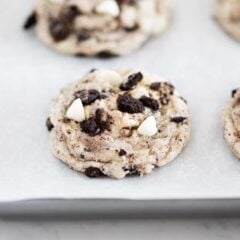 Showing cookies and cream cookies on baking sheet with Oreo crumbs and white chocolate chips on top.