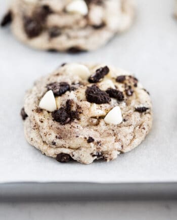 Showing cookies and cream cookies on baking sheet with Oreo crumbs and white chocolate chips on top.